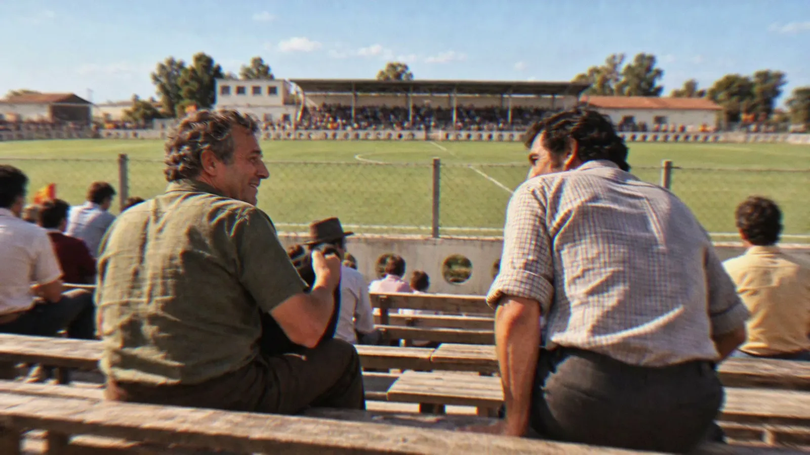 Dois torcedores conversam na arquibancada, com o campo e o pavilhão ao fundo na reconstrução do filme.